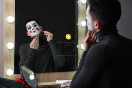Young man applying mime makeup near mirror in dressing room