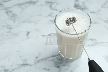 Mini mixer (milk frother) and tasty cappuccino in glass on white marble table, closeup. Space for text
