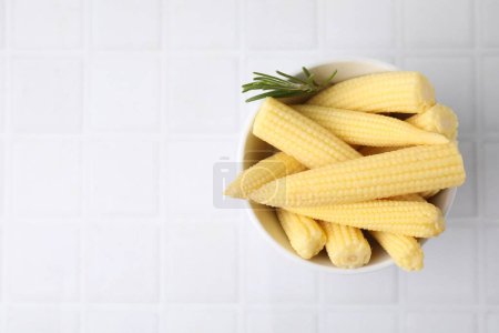 Tasty fresh yellow baby corns in bowl on white tiled table, top view. Space for text