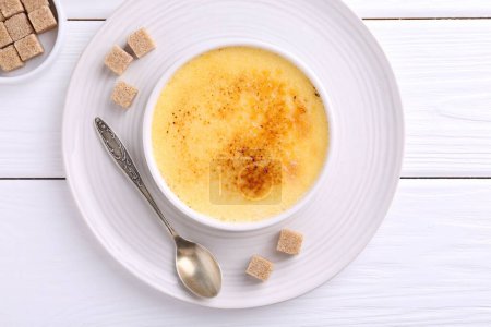 Foto de Deliciosa crema brulee en tazón, cubos de azúcar y cuchara en mesa de madera blanca, vista superior - Imagen libre de derechos
