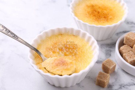 Photo for Delicious creme brulee in bowls, sugar cubes and spoon on white marble table, closeup - Royalty Free Image
