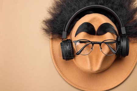 Téléchargez les photos : Visage d'homme en cheveux artificiels, sourcils, lunettes et chapeau sur fond beige, vue de dessus. Espace pour le texte - en image libre de droit