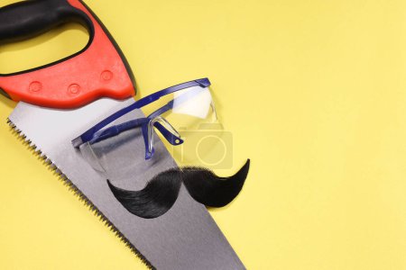 Téléchargez les photos : Visage d'homme fait de moustache artificielle, lunettes de sécurité et scie à main sur fond jaune, vue de dessus. Espace pour le texte - en image libre de droit