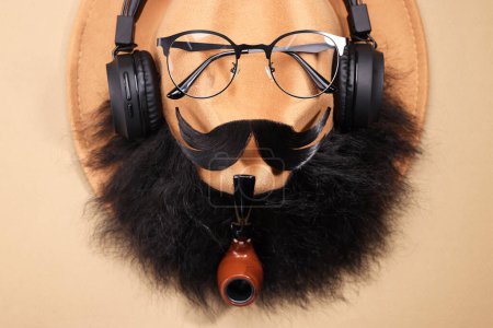 Téléchargez les photos : Visage d'homme fait de moustache artificielle, barbe, lunettes et chapeau sur fond beige, vue de dessus - en image libre de droit