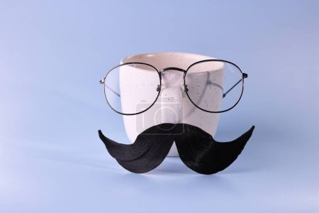 Téléchargez les photos : Visage d'homme fait de moustache artificielle, lunettes et tasse sur fond bleu clair - en image libre de droit