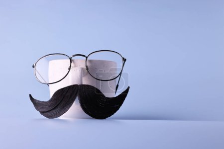 Téléchargez les photos : Visage d'homme fait de moustache artificielle, lunettes et tasse sur fond bleu clair. Espace pour le texte - en image libre de droit
