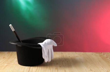Magician's hat, wand and gloves on wooden table against color background, space for text