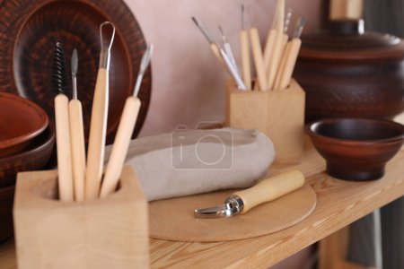 Set of different crafting tools and clay dishes on wooden rack in workshop, closeup
