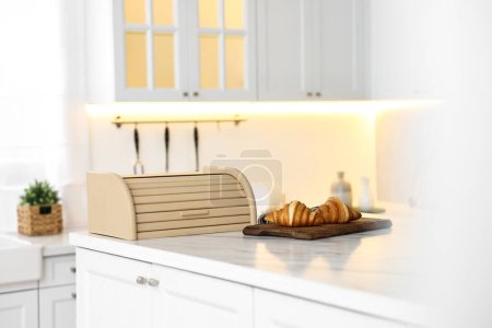 Wooden bread box and board with croissants on white marble countertop in kitchen