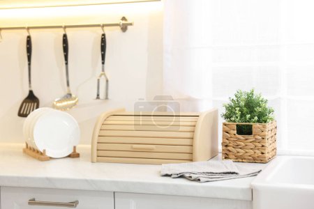 Wooden bread box, houseplant and plates on white marble countertop in kitchen