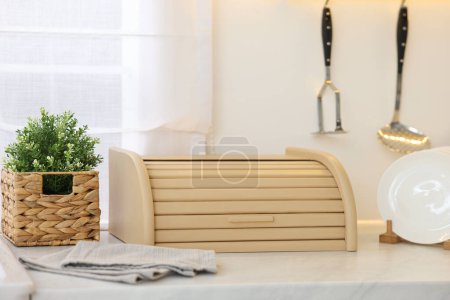 Wooden bread box, houseplant and plates on white marble countertop in kitchen
