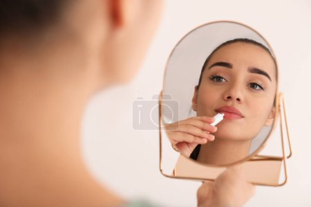 Woman with herpes applying cream on lips in front of mirror against light background