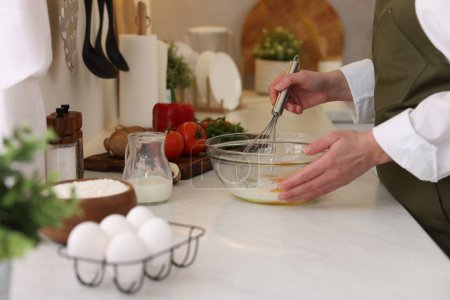Woman whisking eggs in bowl at light table indoors, closeup