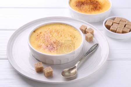 Téléchargez les photos : Délicieuse crème brulée dans des bols, des cubes de sucre et une cuillère sur une table en bois blanc - en image libre de droit