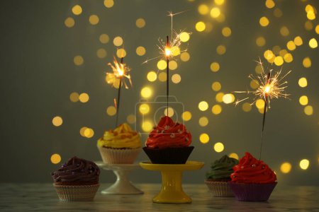 Different colorful cupcakes with sparklers on table against blurred lights