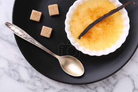 Foto de Deliciosa crema brulee en tazón, vaina de vainilla, cubos de azúcar y cuchara en la mesa de mármol blanco, vista superior - Imagen libre de derechos