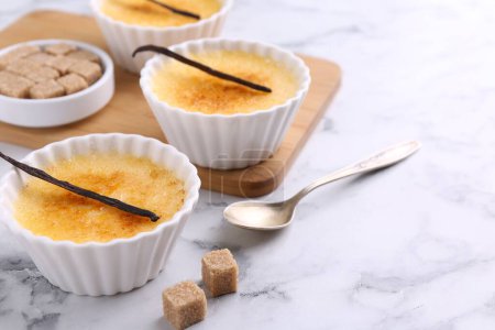 Photo for Delicious creme brulee in bowls, vanilla pods, sugar cubes and spoon on white marble table, closeup. Space for text - Royalty Free Image