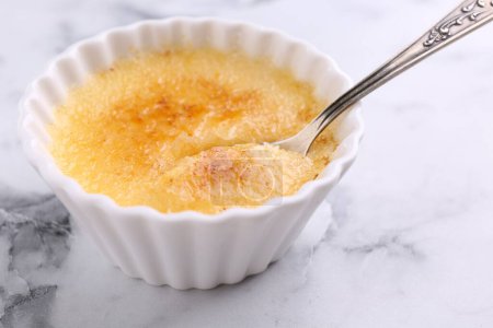 Foto de Deliciosa crema brulee en tazón y cuchara sobre mesa de mármol blanco, primer plano - Imagen libre de derechos