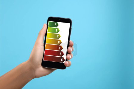 Foto de Calificación de eficiencia energética en la pantalla del teléfono inteligente. Dispositivo de sujeción del hombre sobre fondo azul claro, primer plano - Imagen libre de derechos