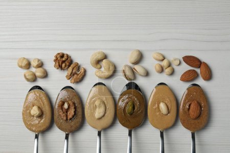 Tasty nut butters in spoons and raw nuts on white wooden table, flat lay. Space for text