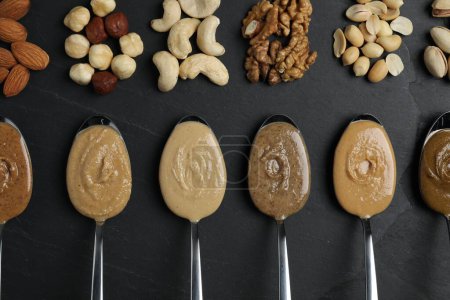 Tasty nut butters in spoons and raw nuts on dark gray textured table, flat lay