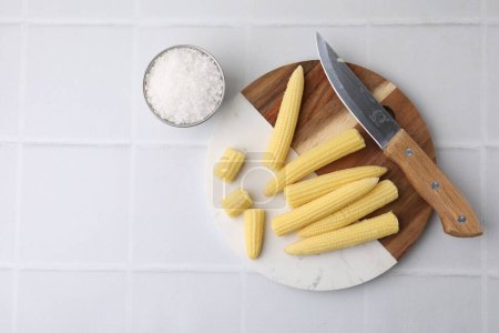 Tasty fresh yellow baby corns and knife on white tiled table, top view. Space for text