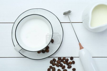 Composición plana con mini batidora (espuma de leche), leche batida y granos de café sobre una mesa de madera blanca