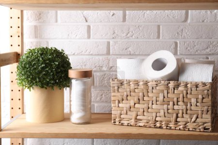 Téléchargez les photos : Rouleaux de papier toilette dans un panier en osier, décor floral et tampons de coton sur une étagère en bois contre un mur de briques blanches - en image libre de droit