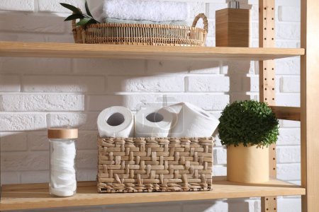 Téléchargez les photos : Rouleaux de papier toilette dans un panier en osier, décor floral et tampons de coton sur une étagère en bois près d'un mur de briques blanches - en image libre de droit