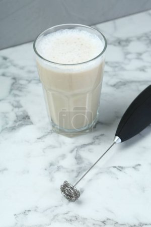 Mini mixer (milk frother) and tasty cappuccino in glass on white marble table