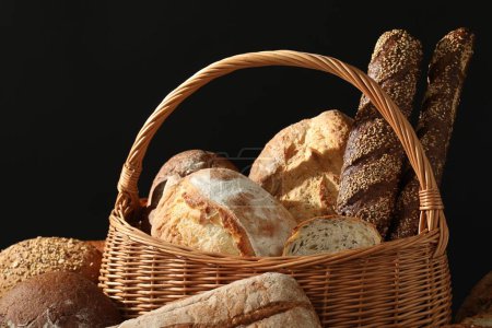 Weidenkorb mit verschiedenen Arten von frischem Brot vor schwarzem Hintergrund