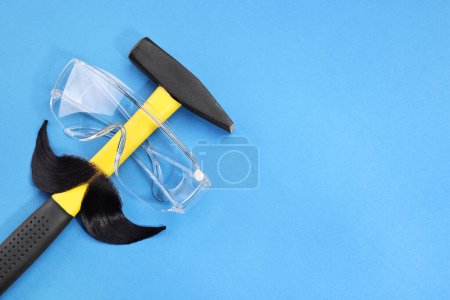 Téléchargez les photos : Visage d'homme fait de moustache artificielle, lunettes de sécurité et marteau sur fond bleu, vue de dessus. Espace pour le texte - en image libre de droit