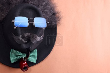Téléchargez les photos : Visage d'homme fait de moustache artificielle, lunettes de soleil et chapeau sur fond brun, vue de dessus. Espace pour le texte - en image libre de droit