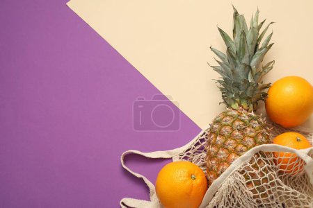 Téléchargez les photos : Sac à ficelle avec ananas frais et oranges sur fond de couleur, plat. Espace pour le texte - en image libre de droit