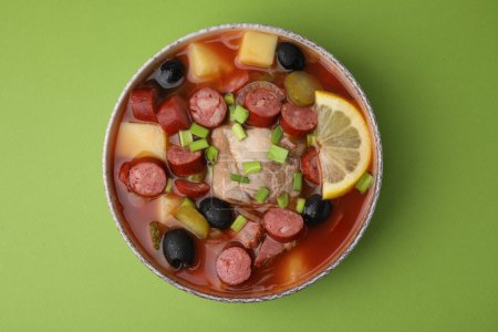 Meat solyanka soup with thin dry smoked sausages in bowl on light green background, top view