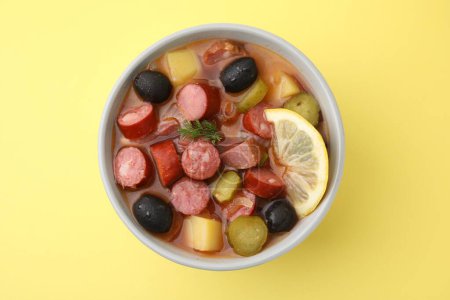 Meat solyanka soup with thin dry smoked sausages in bowl on pale yellow background, top view