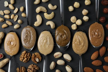 Tasty nut butters in spoons and raw nuts on dark gray table, flat lay