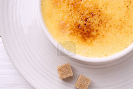 Foto de Deliciosa crema brulee en tazón y terrones de azúcar en la mesa de madera blanca, vista superior - Imagen libre de derechos