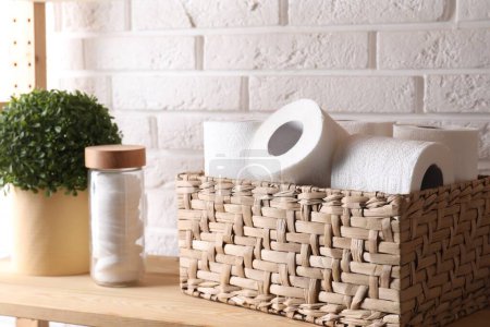 Téléchargez les photos : Rouleaux de papier toilette dans un panier en osier, décor floral et tampons de coton sur une étagère en bois près d'un mur de briques blanches - en image libre de droit