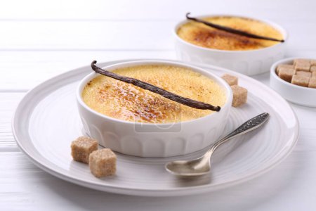 Téléchargez les photos : Délicieuse crème brulée dans des bols, gousses de vanille, cubes de sucre et cuillère sur table en bois blanc, gros plan - en image libre de droit