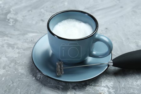 Mini mixer (milk frother) and whipped milk in cup on grey textured table, closeup