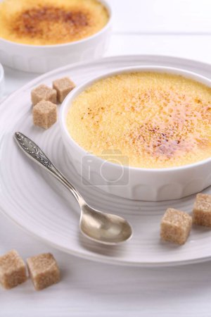 Foto de Deliciosa crema brulee en cuencos, terrones de azúcar y cuchara en mesa de madera blanca - Imagen libre de derechos