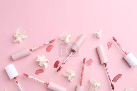 Verschiedene Lippenglanzungen, Applikatoren und Blumen auf rosa Hintergrund, flach liegend. Raum für Text