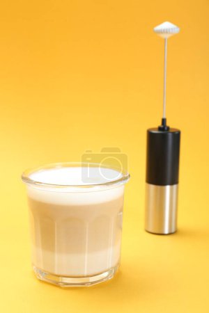 Mini mixer (milk frother) and tasty cappuccino in glass on yellow background