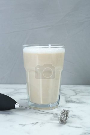 Mini mixer (milk frother) and tasty cappuccino in glass on white marble table