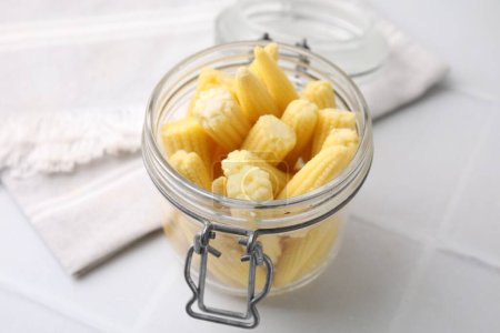 Tasty fresh yellow baby corns in glass jar on white tiled table, closeup