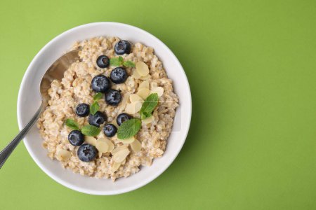 Tasty oatmeal with blueberries, mint and almond petals in bowl on light green background, top view. Space for text