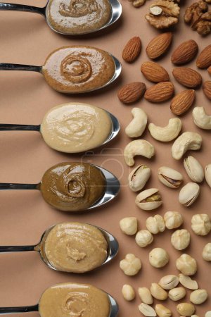 Tasty nut butters in spoons and raw nuts on light brown table, flat lay