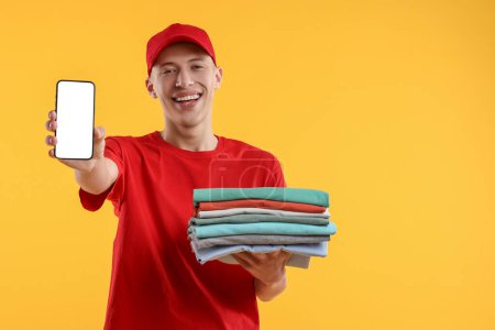 Photo for Dry-cleaning delivery. Happy courier holding folded clothes and smartphone on orange background, space for text - Royalty Free Image