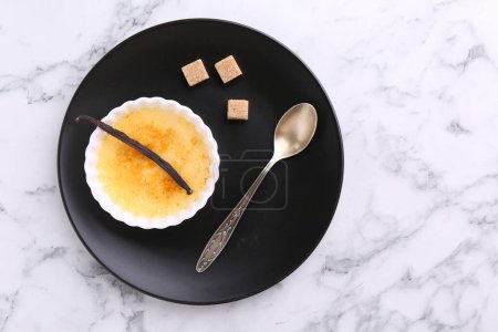 Téléchargez les photos : Délicieuse crème brulée dans un bol, gousse de vanille, cubes de sucre et cuillère sur table en marbre blanc, vue sur le dessus. Espace pour le texte - en image libre de droit